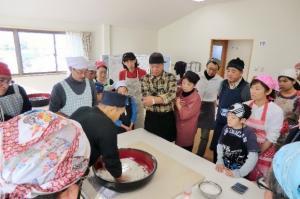エプロンや三角巾を付けた多くの参加者が、そば打ちを行っている男性の作業の様子を見ている写真