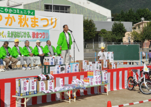 イベントステージ上に設置された椅子に座る黄緑色の法被を着た方々、スタンドマイク前に立ち話をしている男性、ステージの前に沢山の景品が並べられている写真