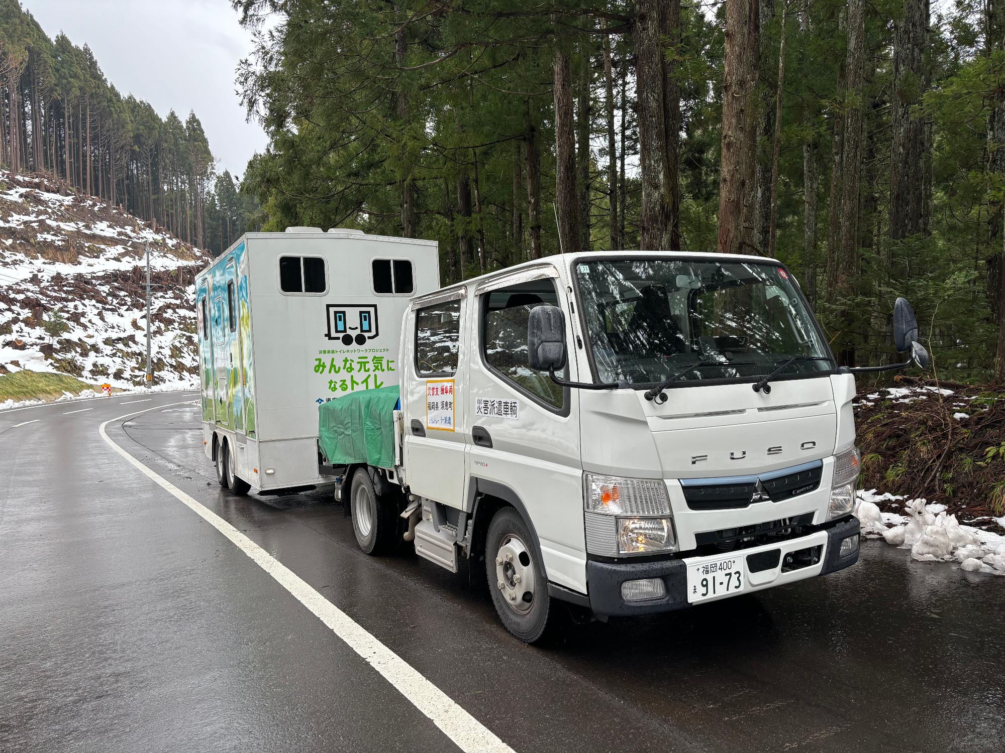 雪が残る山道を走る牽引車とトイレトレーラー