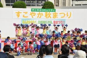 イベント用ステージで黄色やピンク色や水色の衣装を着てハンドフリーチアポンポンを持った子供たちが踊っている写真