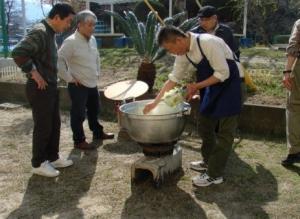 エプロンをつけた男性が大きな鍋の中に材料を入れており、その調理の様子をみている方々の写真