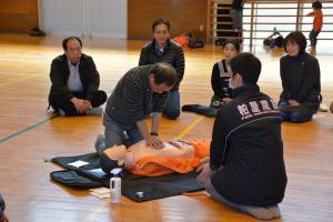 心肺蘇生訓練人形を使い救命講習を受けている男性とその様子を見ている参加者の写真