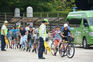 縦一列になって子供たちが自転車に乗っており、黄色い旗を持ち、黄色い帽子とベストを着た男性に自転車の乗り方について指導を受けている自転車教室の様子の写真